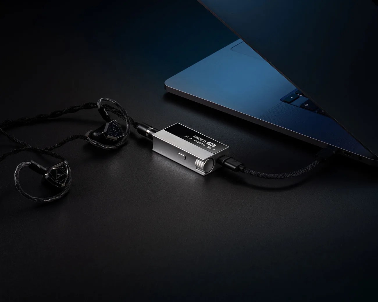 Silver rectangular digital-to-analog converter connected to black earphones and a laptop on a dark surface.