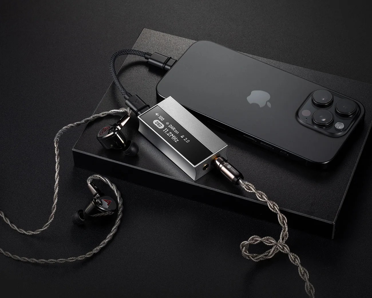 Black iPhone with triple camera lenses connected to a silver digital audio player and black in-ear monitors with braided cables on a black surface.