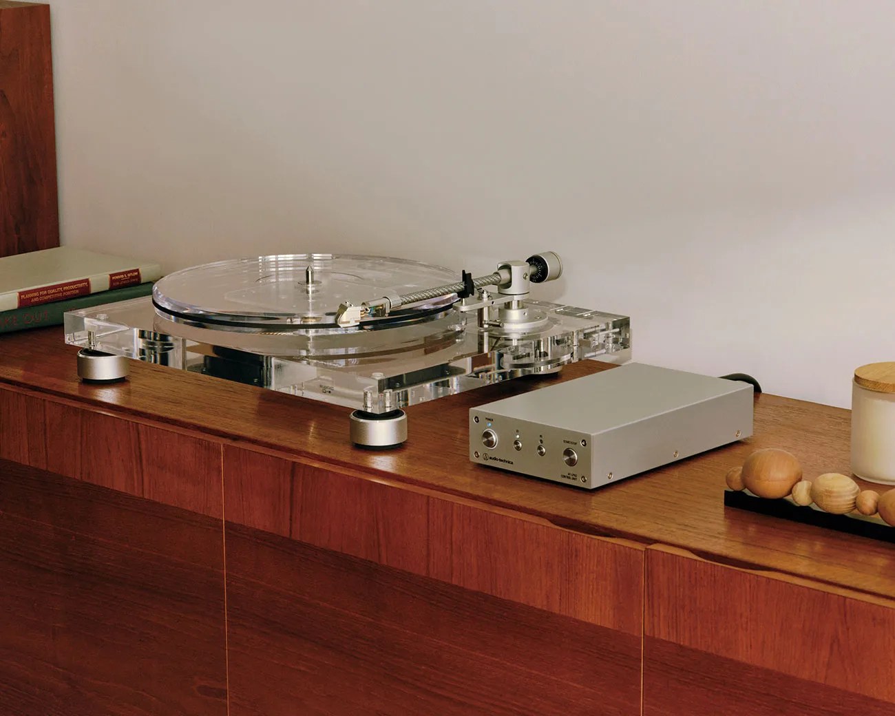 Clear acrylic turntable with silver tonearm and a silver audio control box on a wooden cabinet.