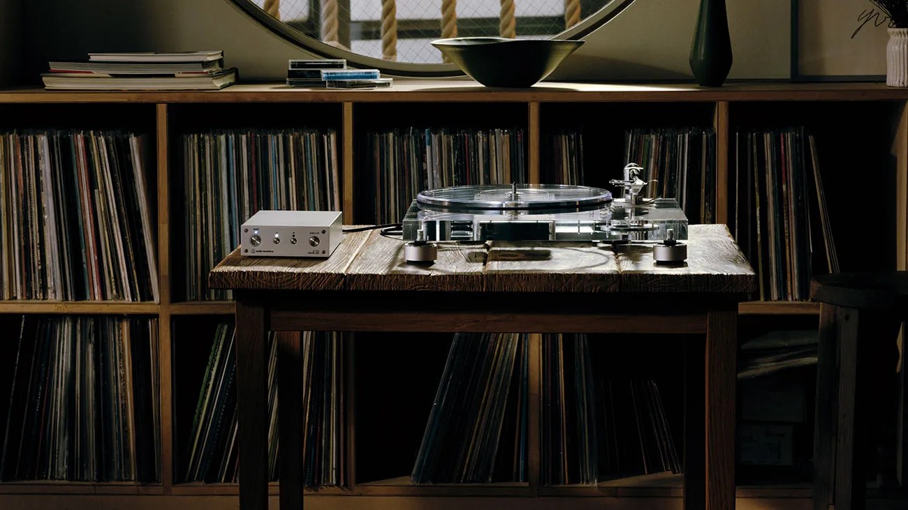 Clear acrylic turntable with silver control box on a wooden table in front of a wooden shelf filled with vinyl records.
