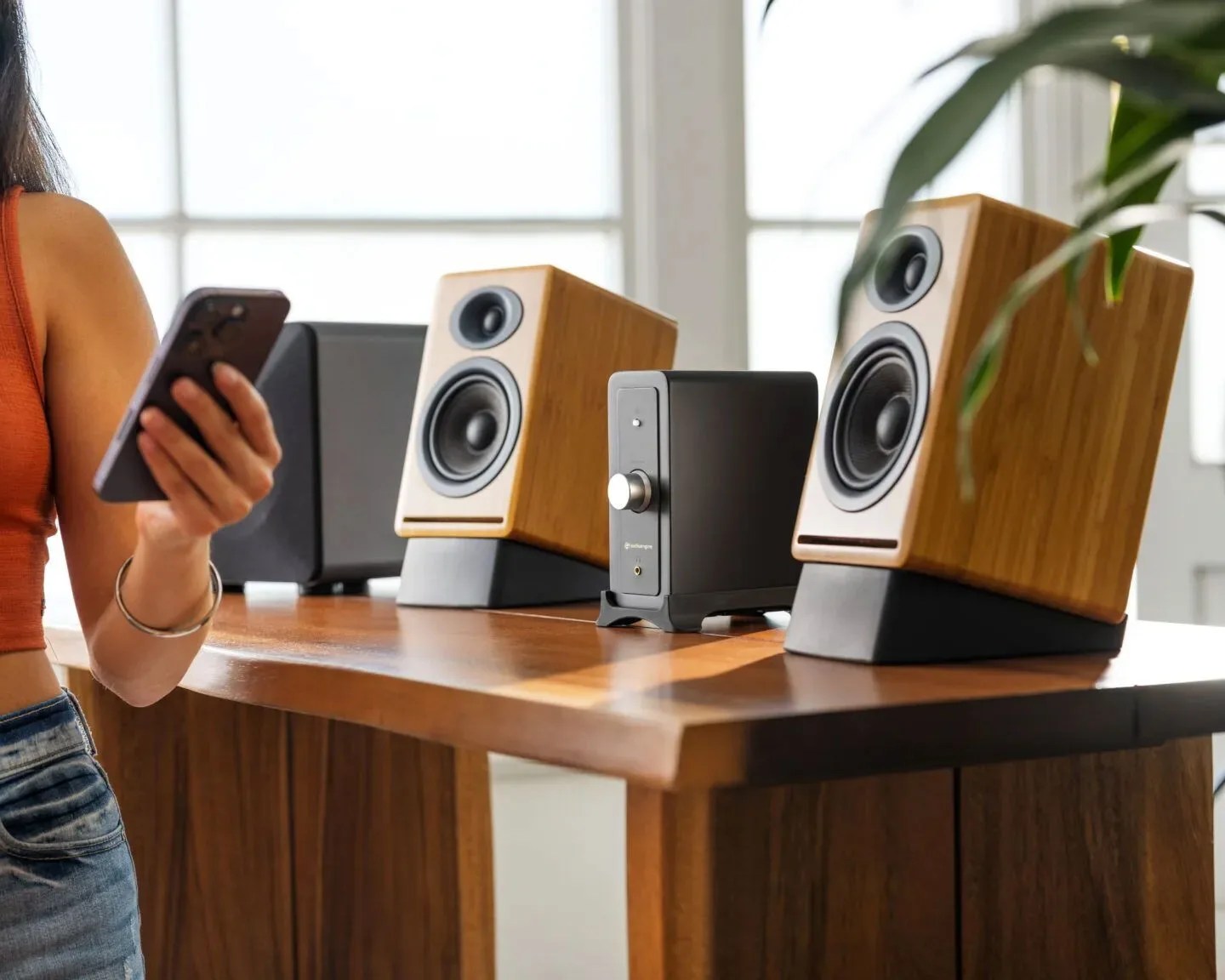 Pair of wooden bookshelf speakers and a black amplifier on a wooden table with a person holding a smartphone nearby.