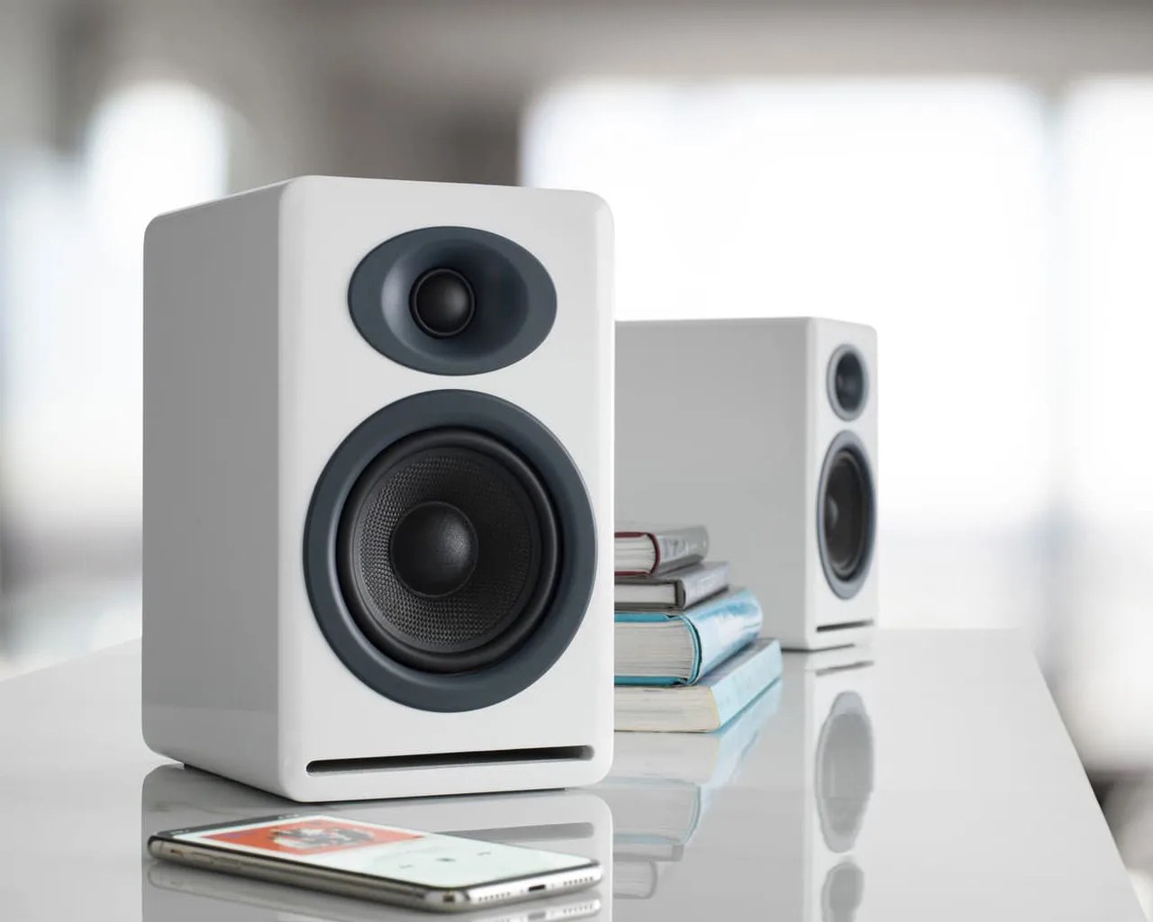 Pair of white bookshelf speakers with black drivers on a glossy white surface next to stacked books and a smartphone.