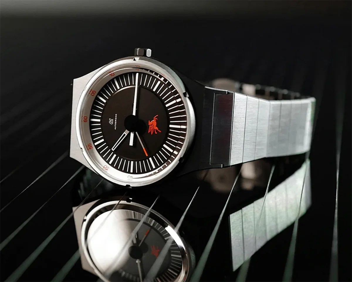 Silver metal wristwatch with black dial, white minute markers, and a red logo, reflected on a glossy black surface.