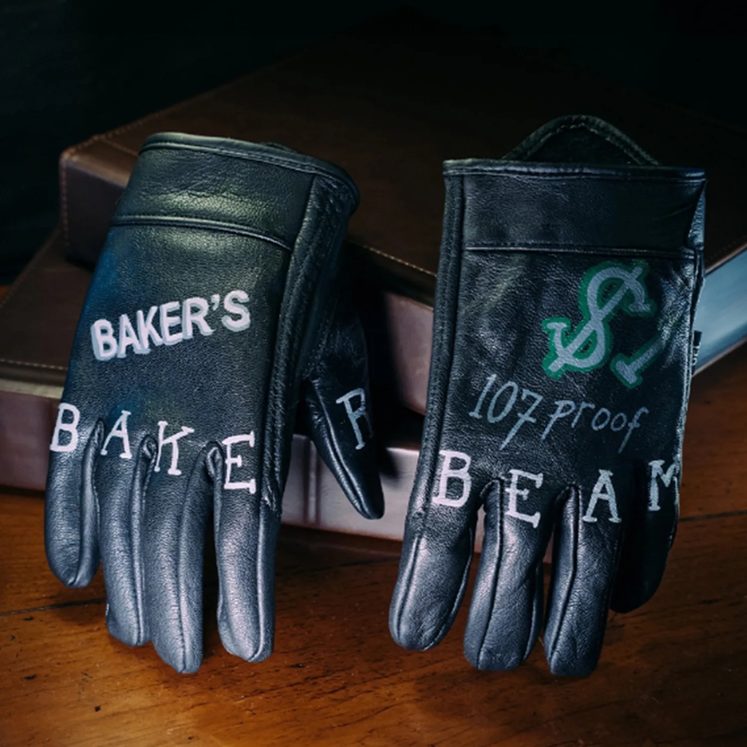Pair of black leather gloves with white and green text reading "Baker's," "107 proof," and "Beam" on a wooden surface.