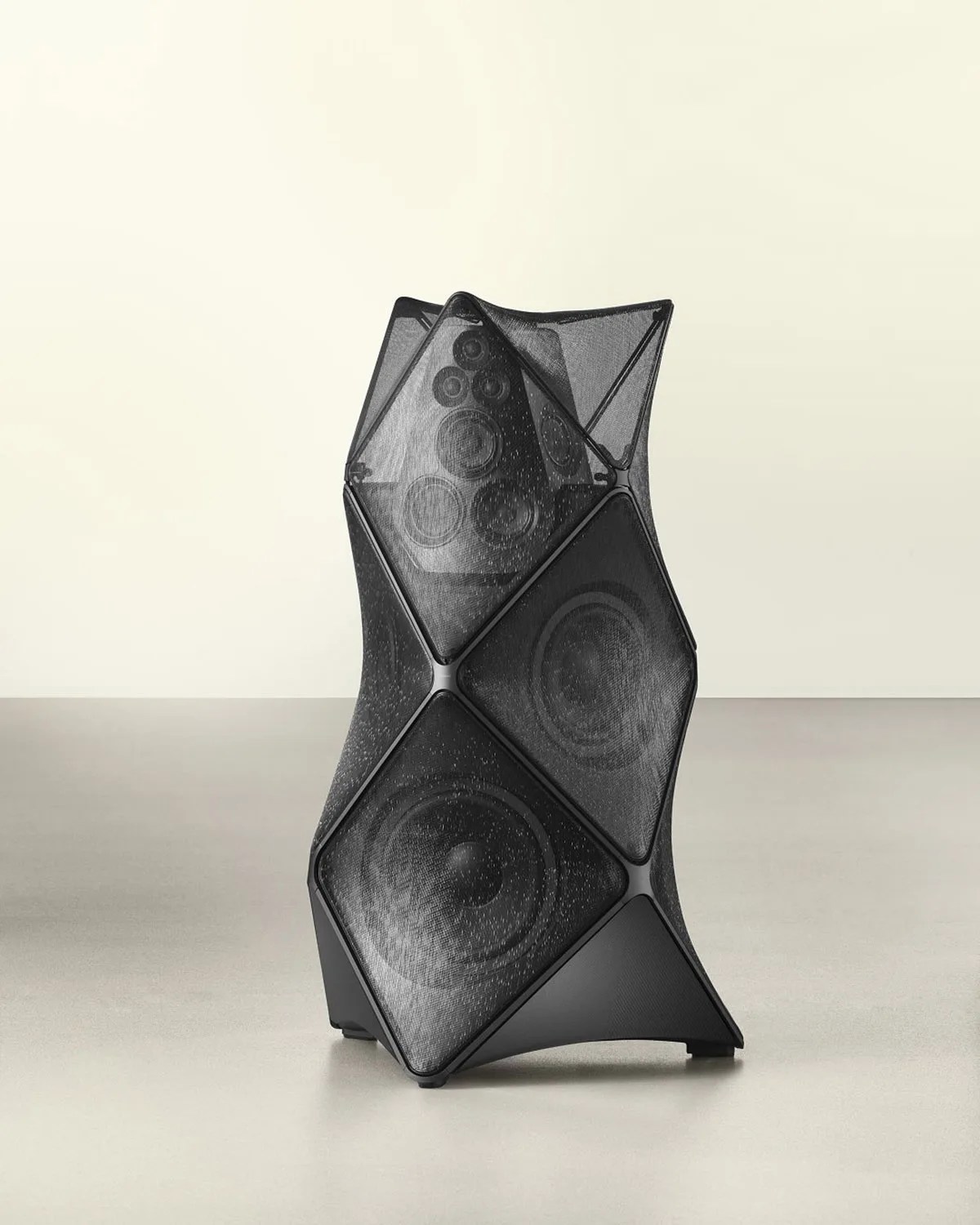 Black modern speaker with a geometric mesh cover and multiple visible drivers on a light floor.