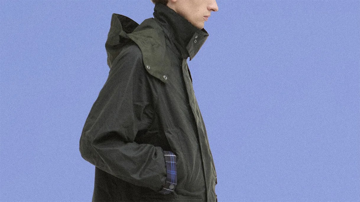 Black hooded jacket with high collar and blue plaid shirt cuffs worn by a person against a blue background.