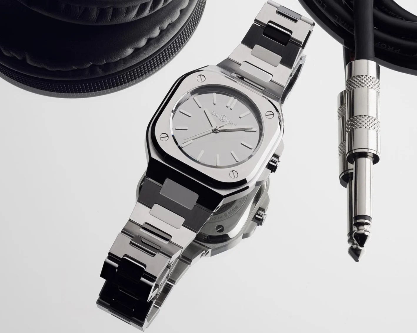 Polished stainless steel wristwatch with a square case and minimalist dial, placed on a reflective surface near black headphones and audio cables.