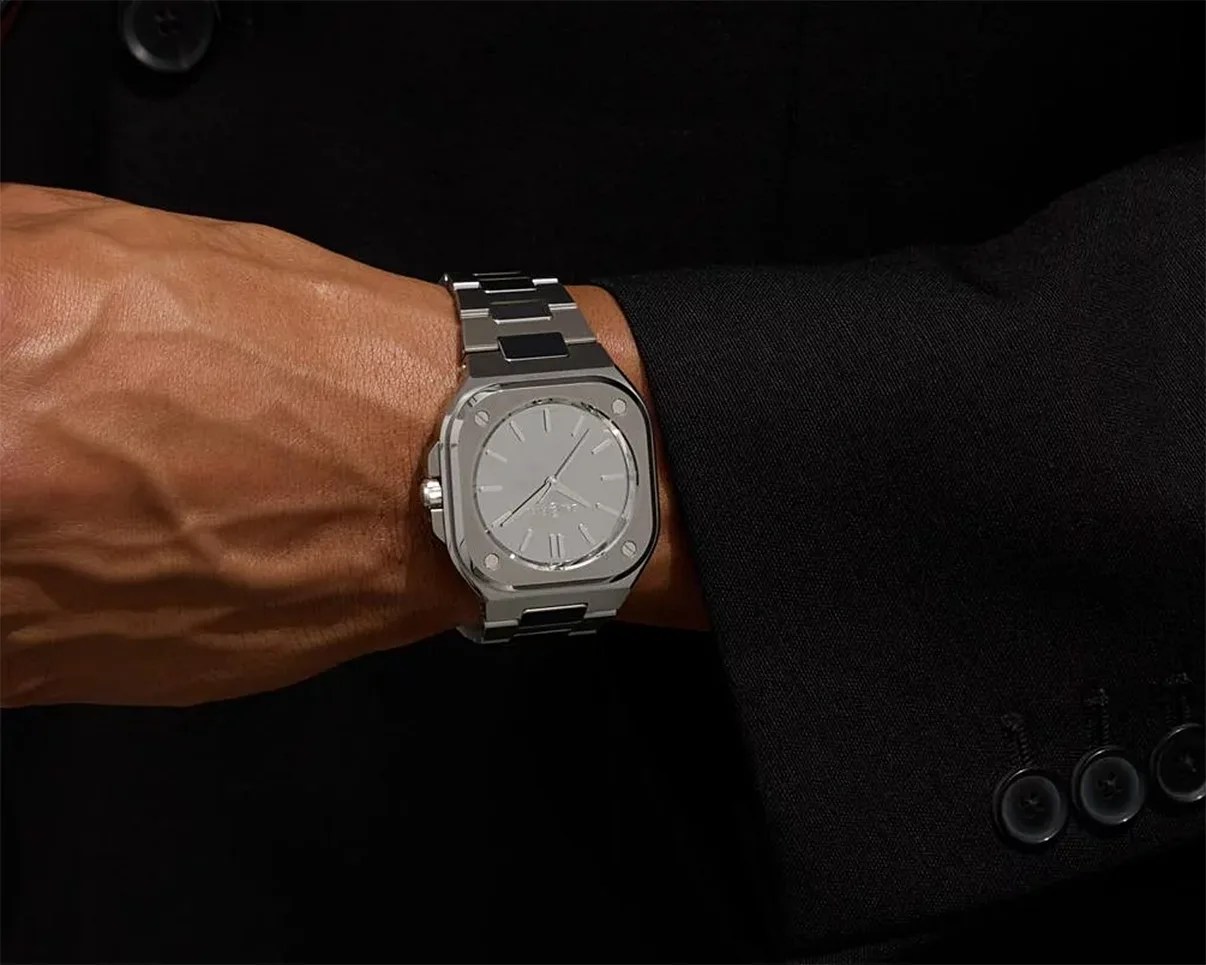 Silver metal wristwatch with a square face and black accents worn on a wrist with a black suit sleeve.
