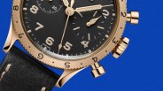 Close-up of a rose gold chronograph watch with black dial and black leather strap on a blue background.