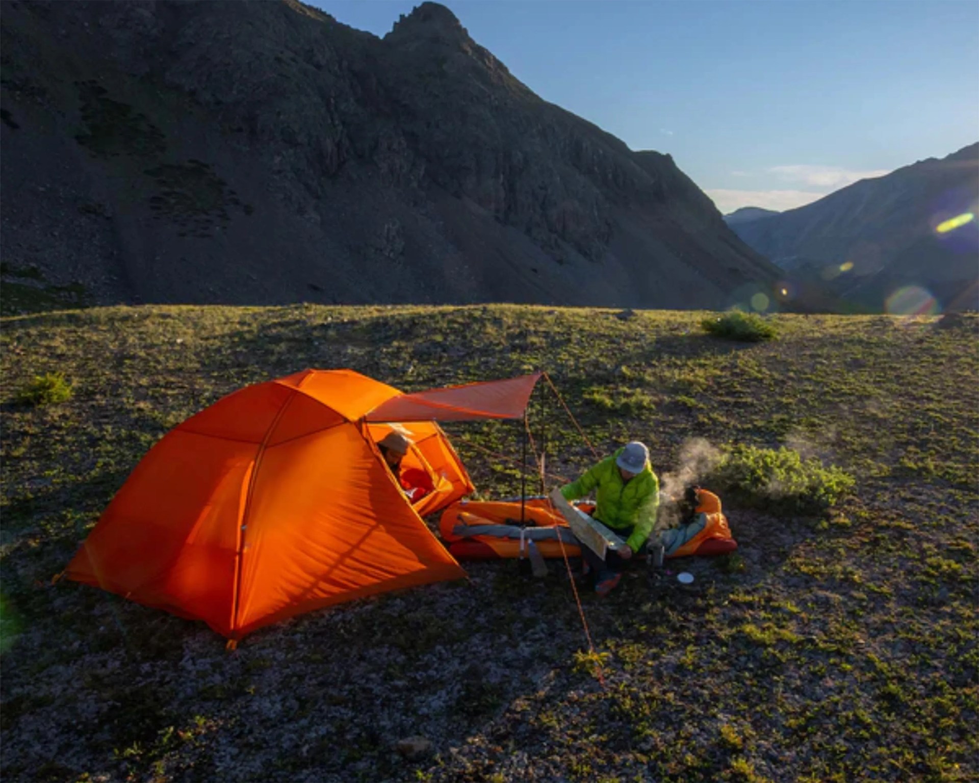 Orange camping tent pitched on rocky terrain with two people, one inside the tent and one sitting outside with a map.