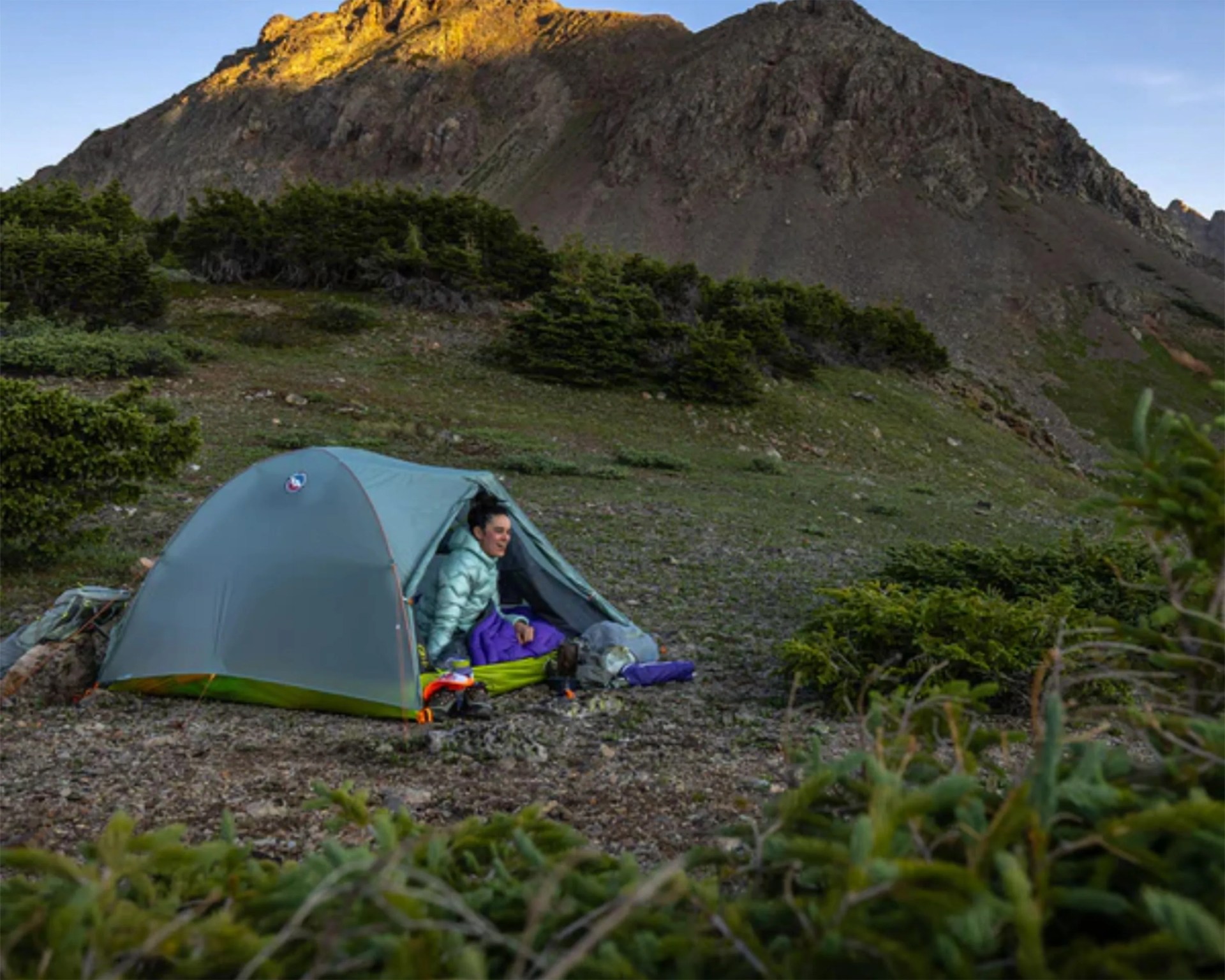 Light blue dome tent pitched on rocky ground with a person sitting inside near green shrubs and a mountain.