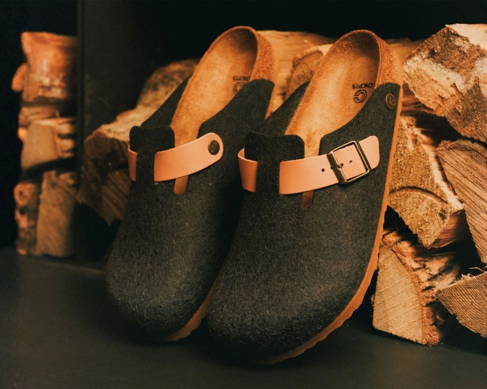 Pair of dark gray felt clogs with tan leather straps and buckles placed near stacked firewood.