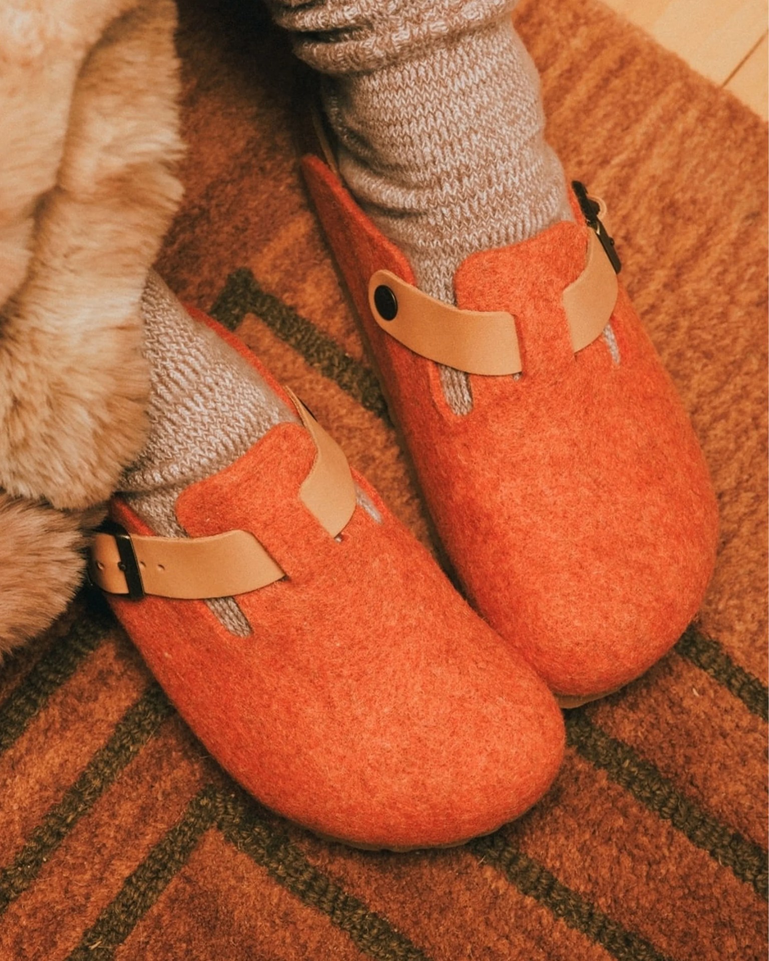 Orange felt clogs with tan leather straps worn with light gray knit socks on a brown patterned rug.