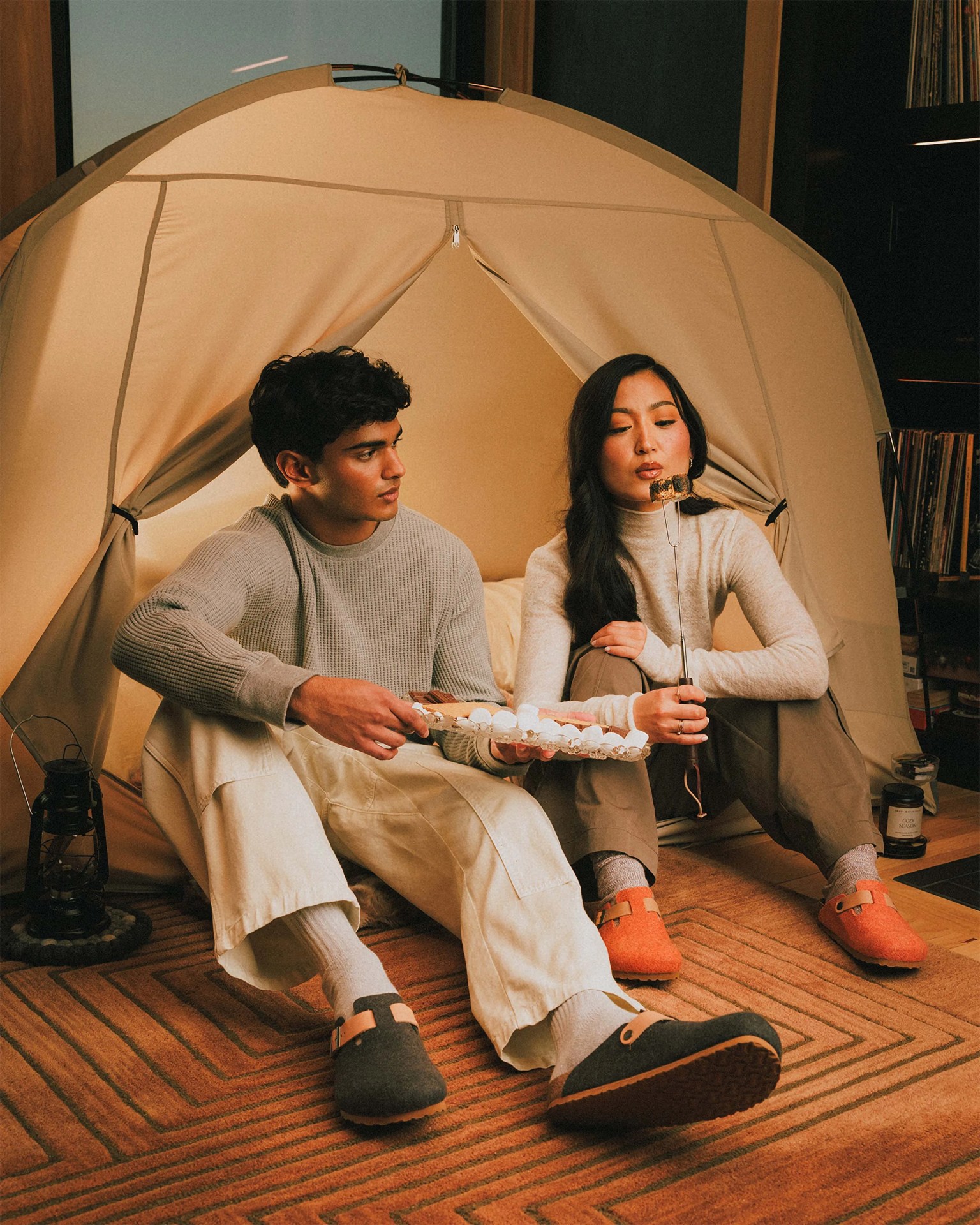 Two people sitting on a brown patterned rug in front of a beige tent, roasting marshmallows, wearing wool slip-on shoes in dark gray and orange.