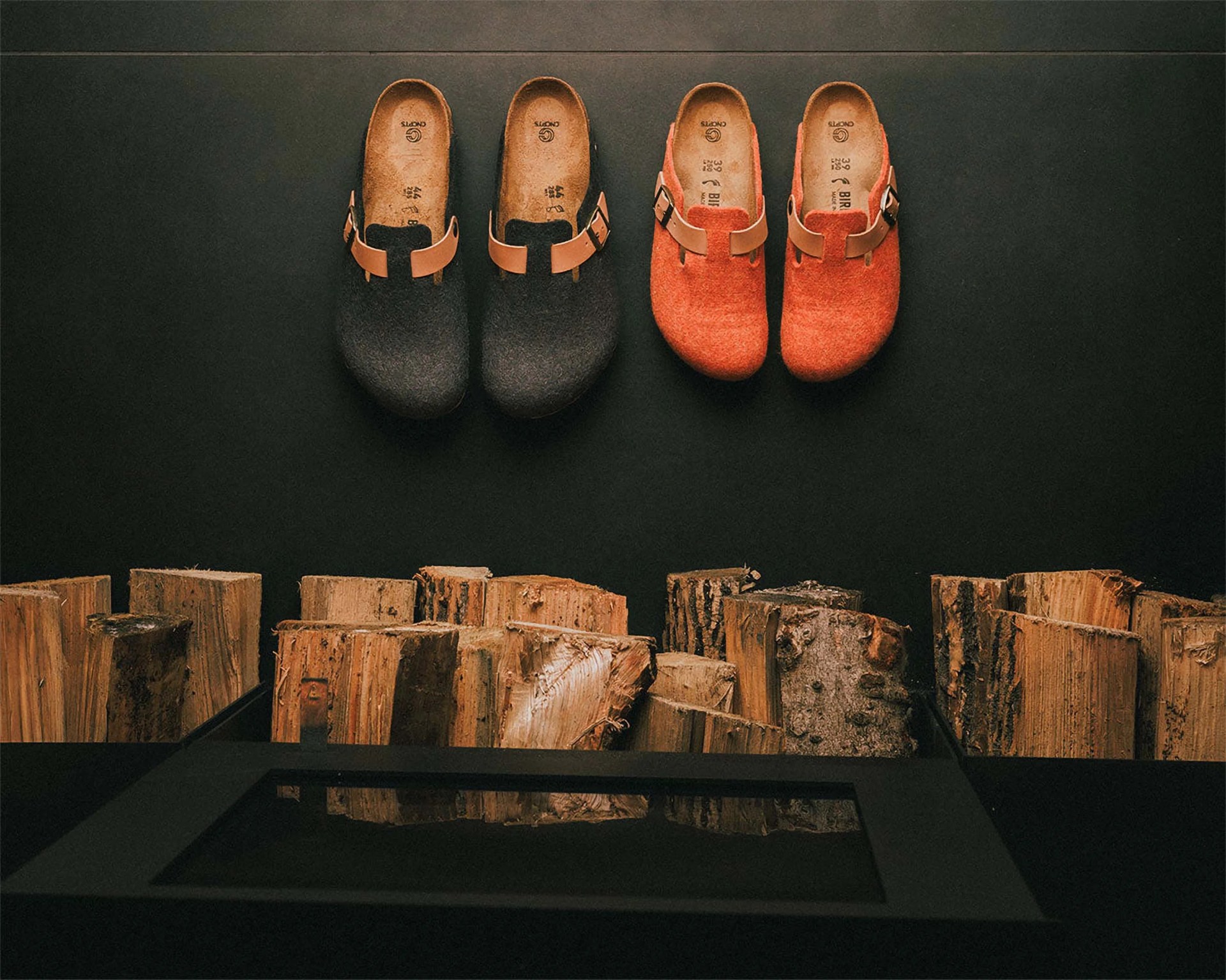 Two pairs of Birkenstock clogs, one black and one orange, displayed on a black wall above stacked firewood.
