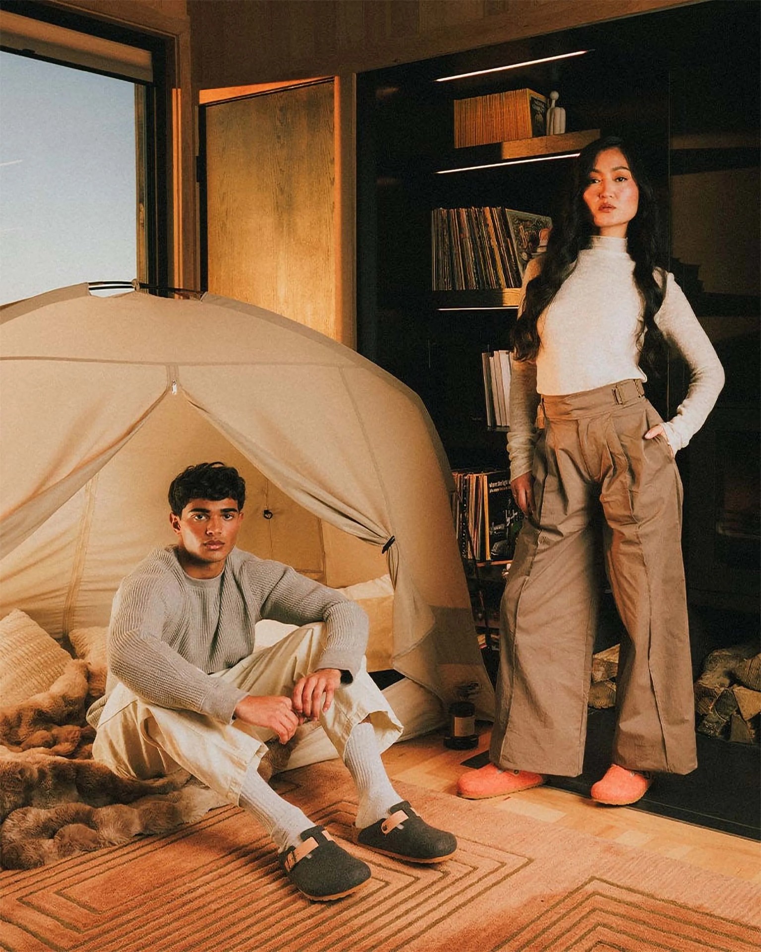 Man sitting inside a beige indoor tent wearing a gray sweater, cream pants, white socks, and black slip-on shoes, and woman standing nearby in a cream turtleneck, wide-legged beige pants, and orange slippers.