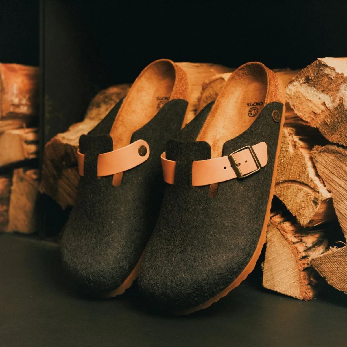 Pair of dark gray felt clogs with tan leather straps and buckles, placed near stacked firewood.