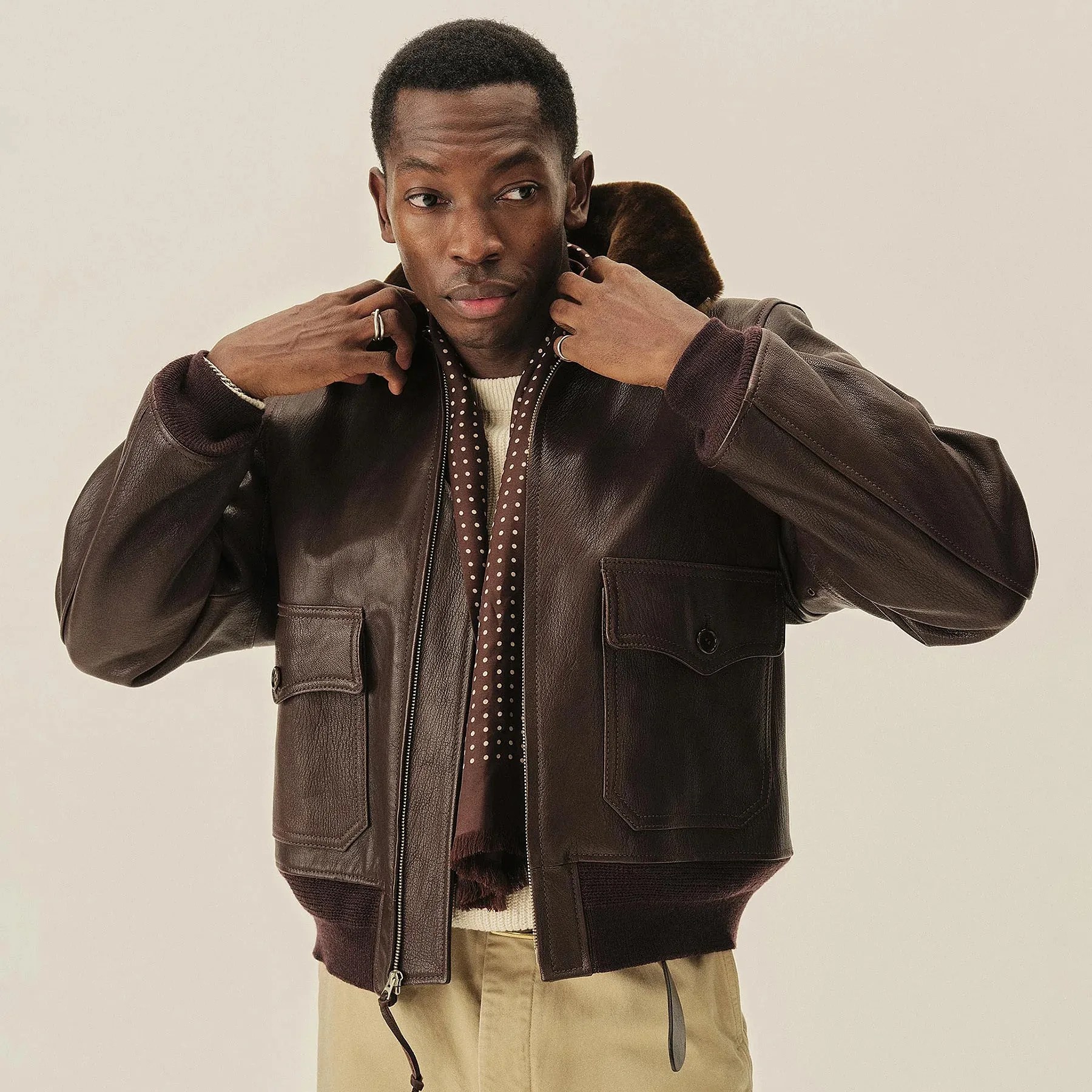 Man wearing a dark brown leather bomber jacket with large front pockets and a fur collar, paired with beige pants.