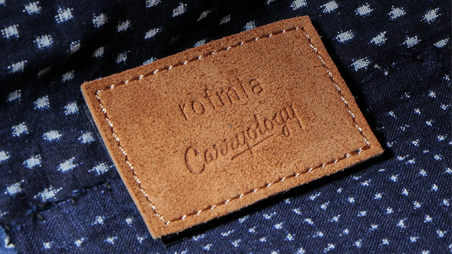 Brown suede rectangular patch with white stitching and embossed text "roimla Cowology" on dark blue fabric with white dots.