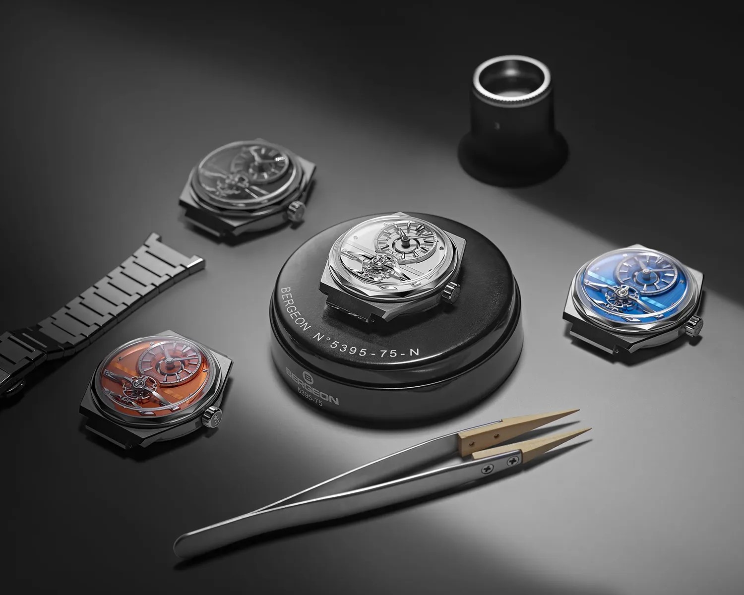 Four mechanical watch movements with different colored dials (orange, silver, blue, and gray) arranged around a Bergeon watchmaker's tool and a pair of tweezers on a gray surface.