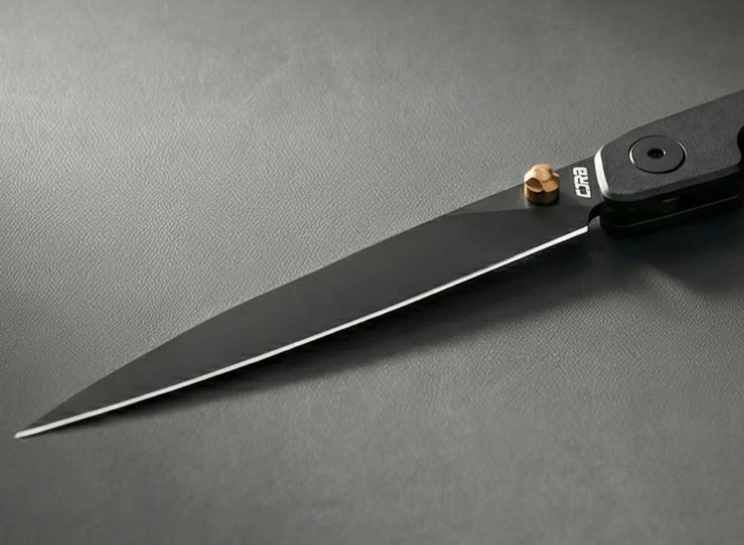 Black folding knife with a sharp pointed blade and a gold thumb stud on a gray textured surface.