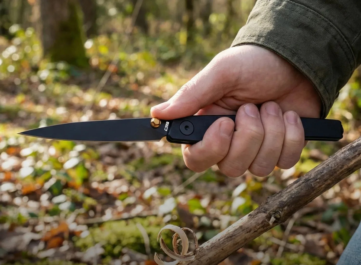 Hand holding a black folding knife with a gold pivot cutting a wood shaving from a stick outdoors.