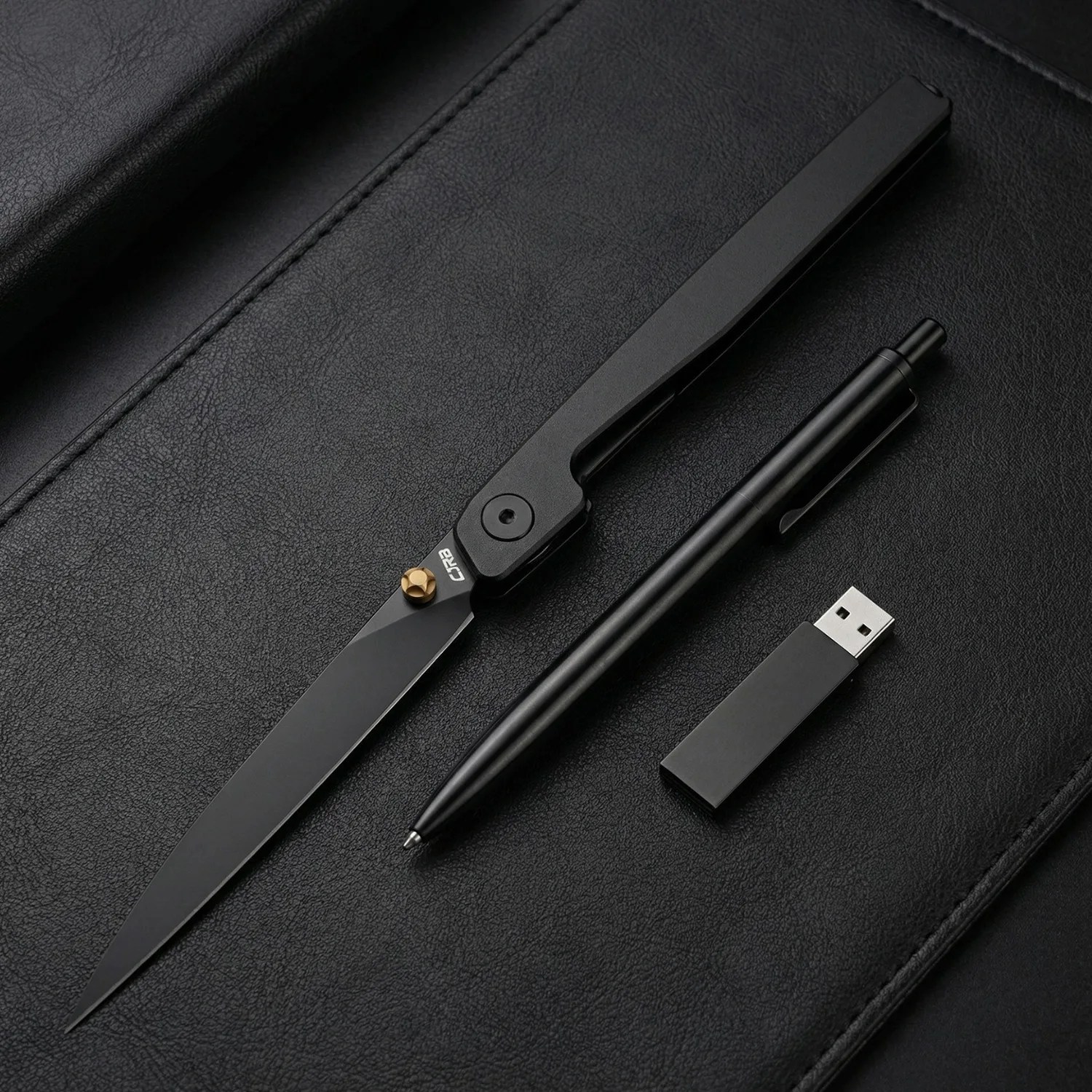 Black folding knife with gold pivot, black click pen, and black USB flash drive on black textured surface.