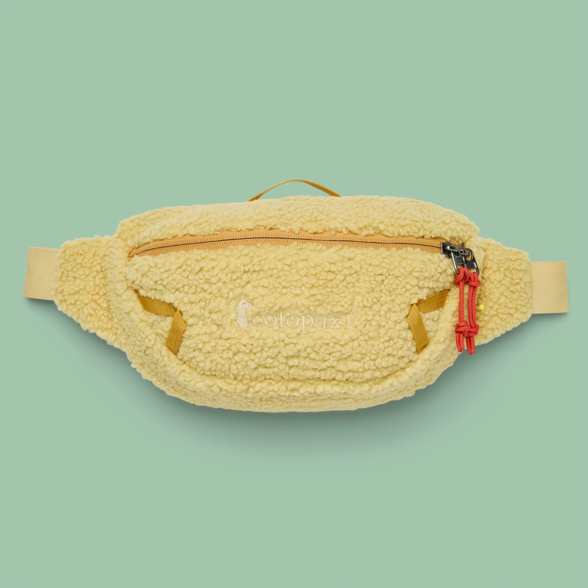 Yellow sherpa fleece fanny pack with front zipper and red zipper pulls on a green background.