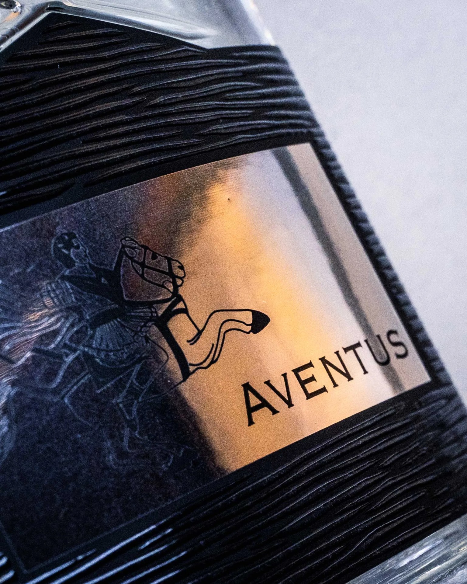 Close-up of a black textured bottle with a metallic label featuring a horse and rider illustration and the word "AVENTUS.
