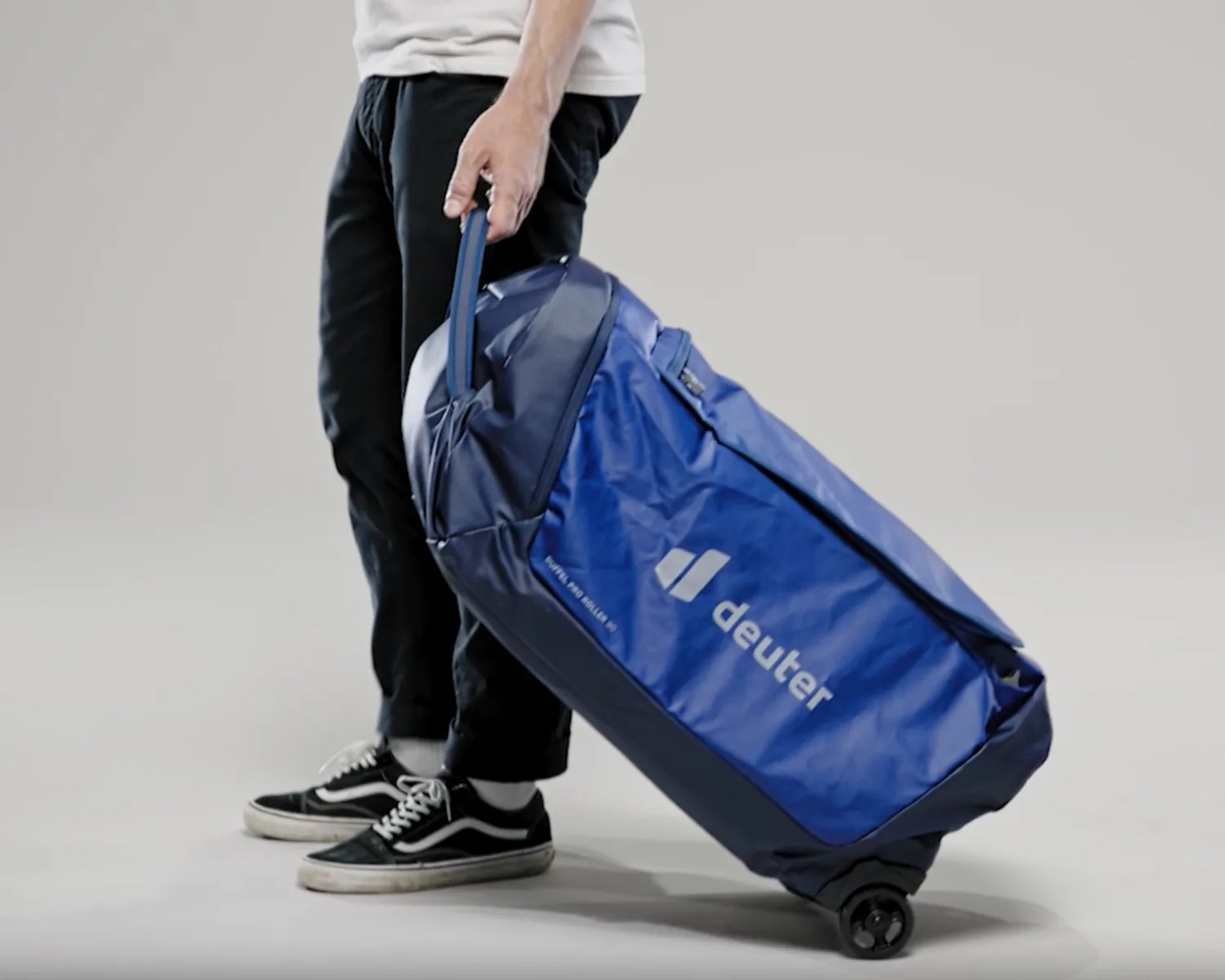 Person holding a blue Deuter wheeled duffel bag with black wheels on a gray background.