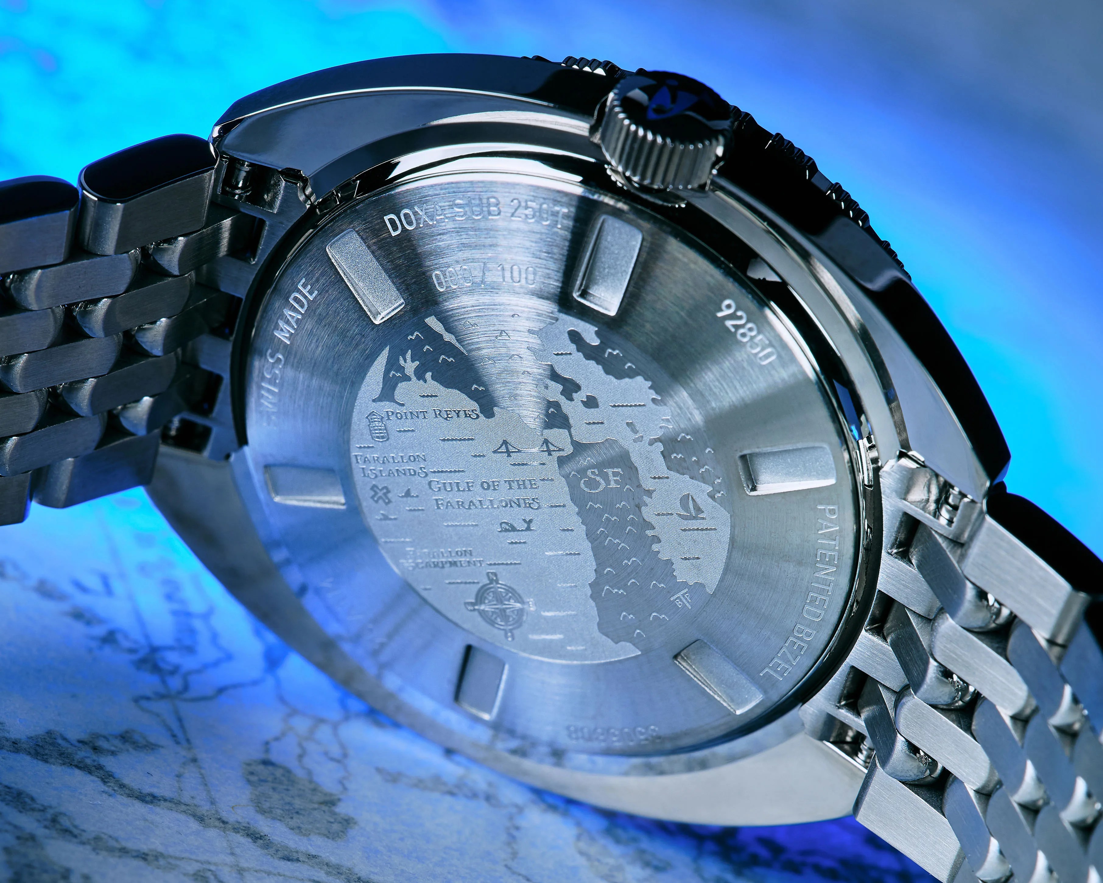 Back of a stainless steel Doxa Sub 250T watch with an engraved map and metal bracelet under blue light.