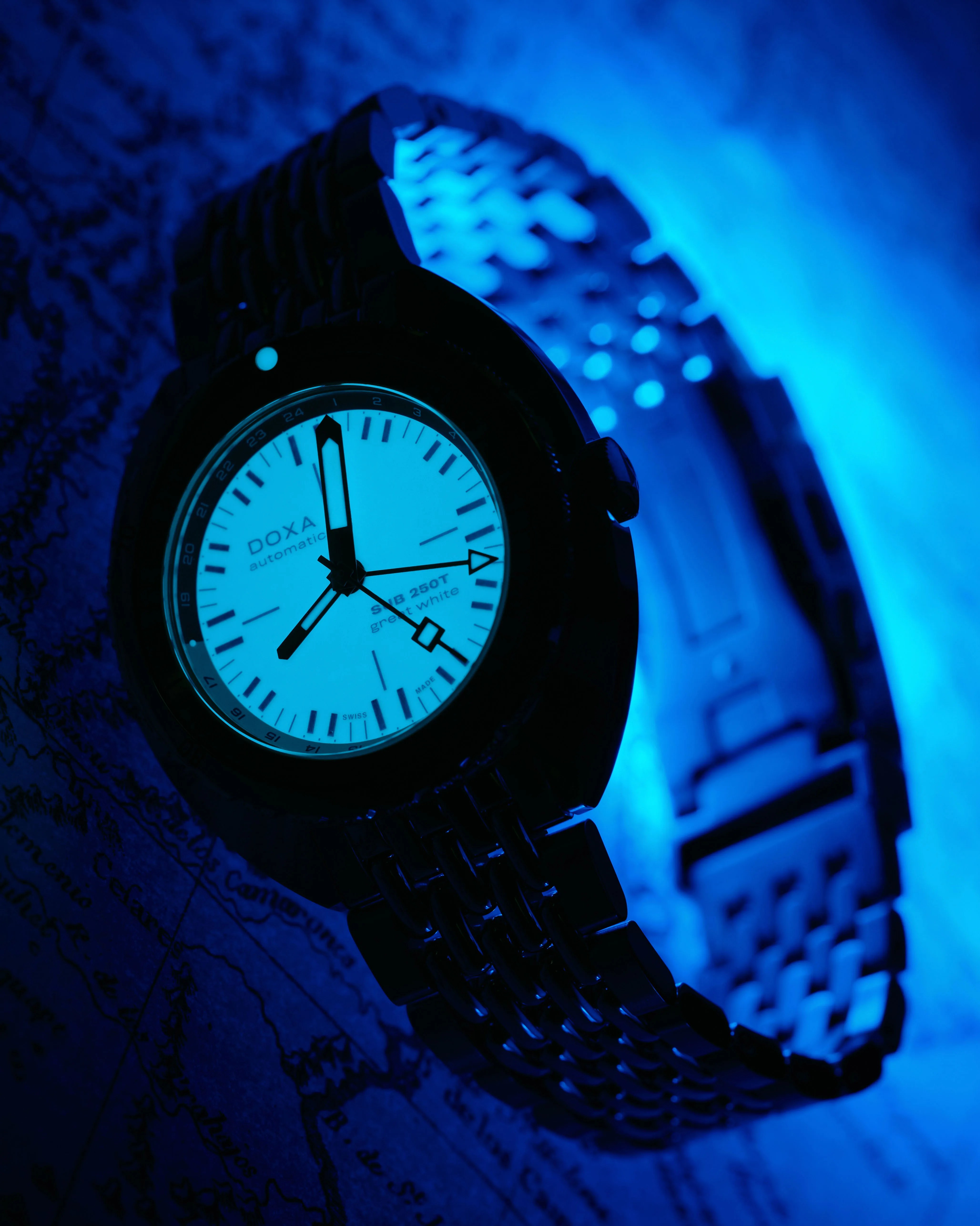 Black metal bracelet watch with glowing white dial and black hands under blue light.