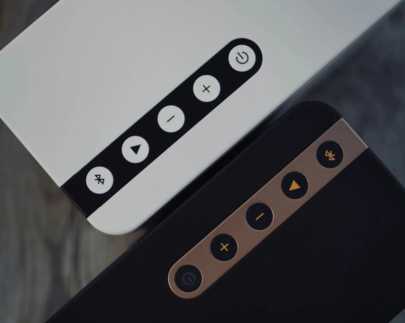 Close-up of two rectangular Bluetooth speakers, one white with black buttons and one black with bronze buttons, showing power, play, volume, and Bluetooth icons.