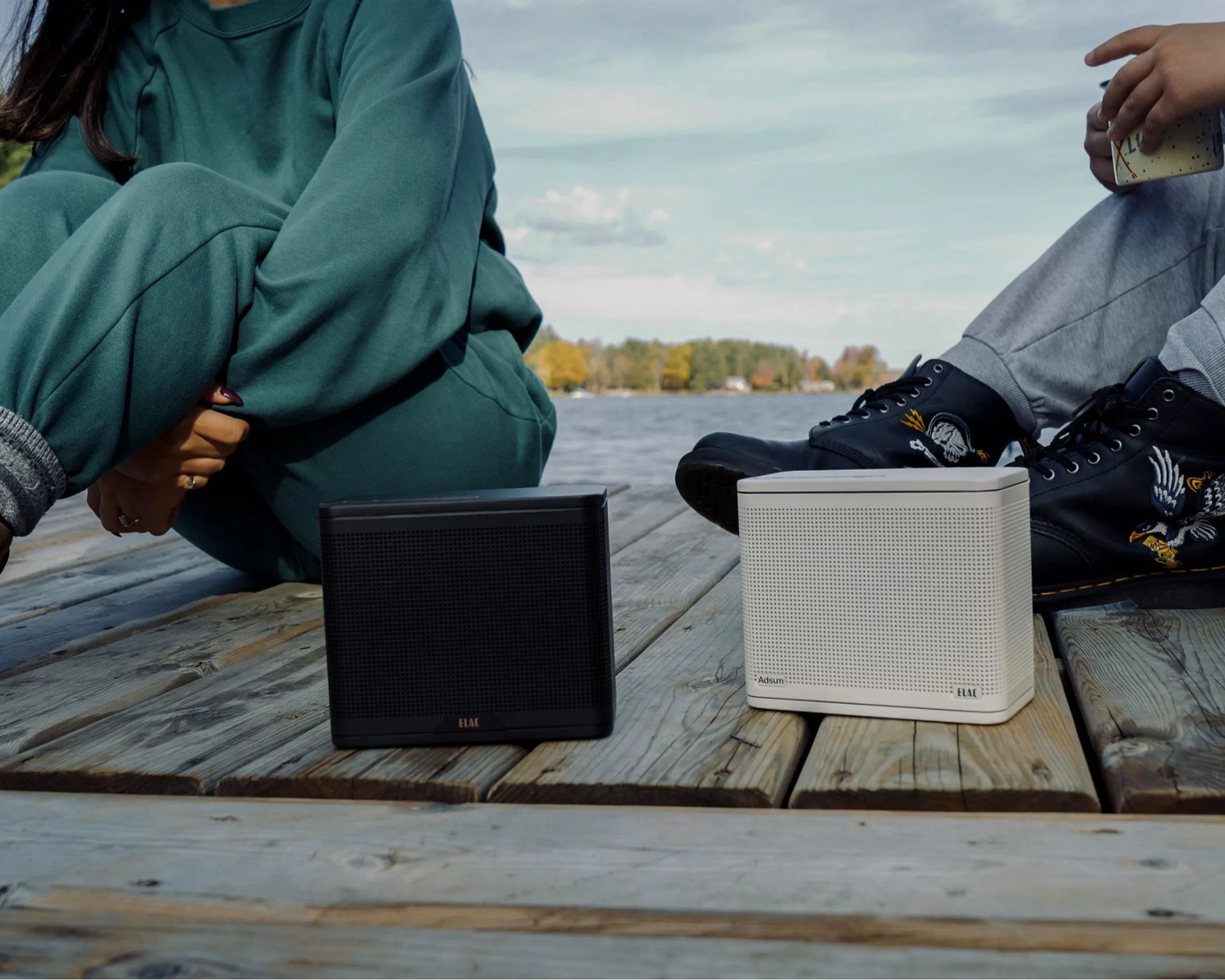 Two rectangular wireless speakers, one black and one white, placed on a wooden dock near a body of water with people sitting nearby.