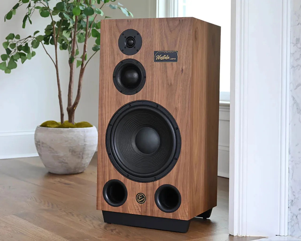 Wood-finished Nostalgia Center speaker with three drivers and two bass ports on a wooden floor near a plant.