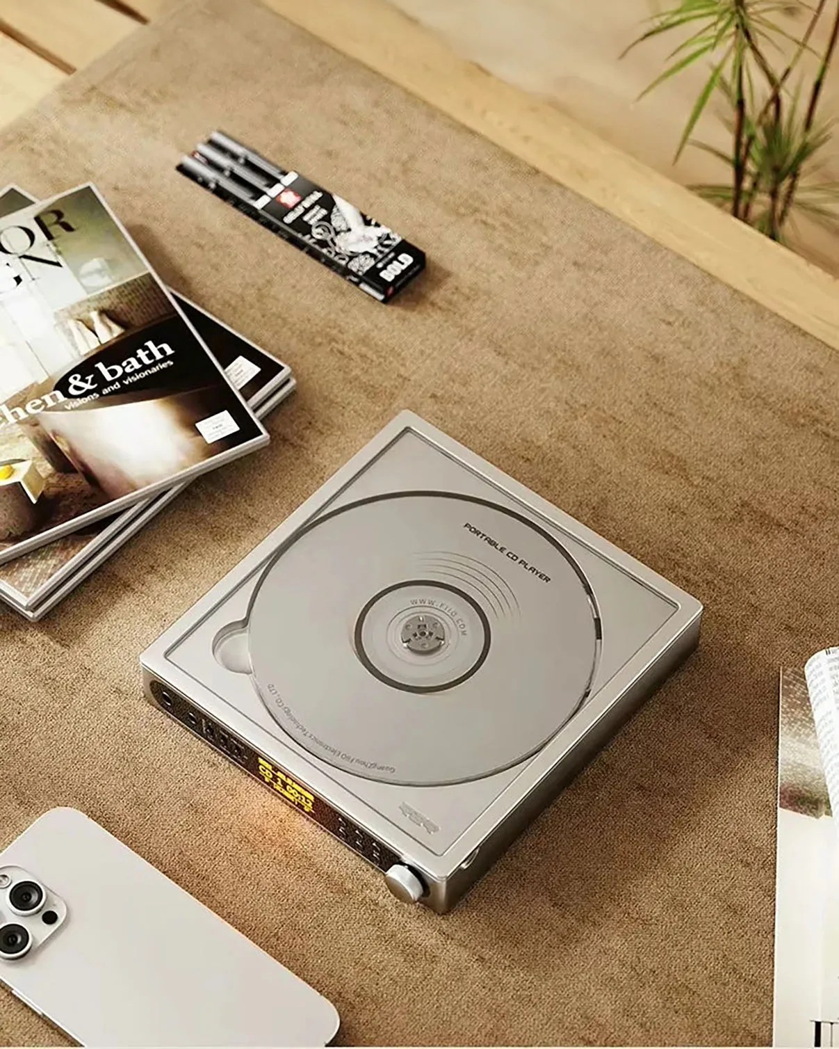 Silver portable CD player with a CD inside on a brown fabric surface surrounded by magazines and a smartphone.