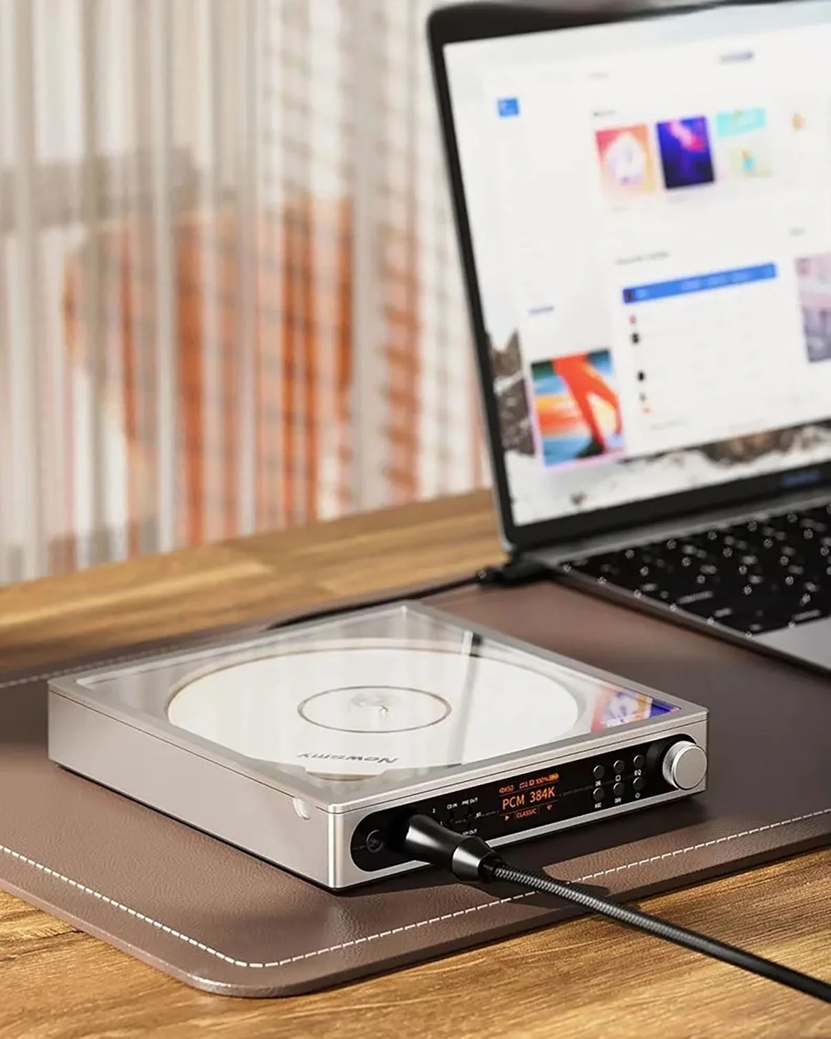 Silver digital audio player with a clear top cover and a connected black cable, placed on a brown mat next to a laptop.