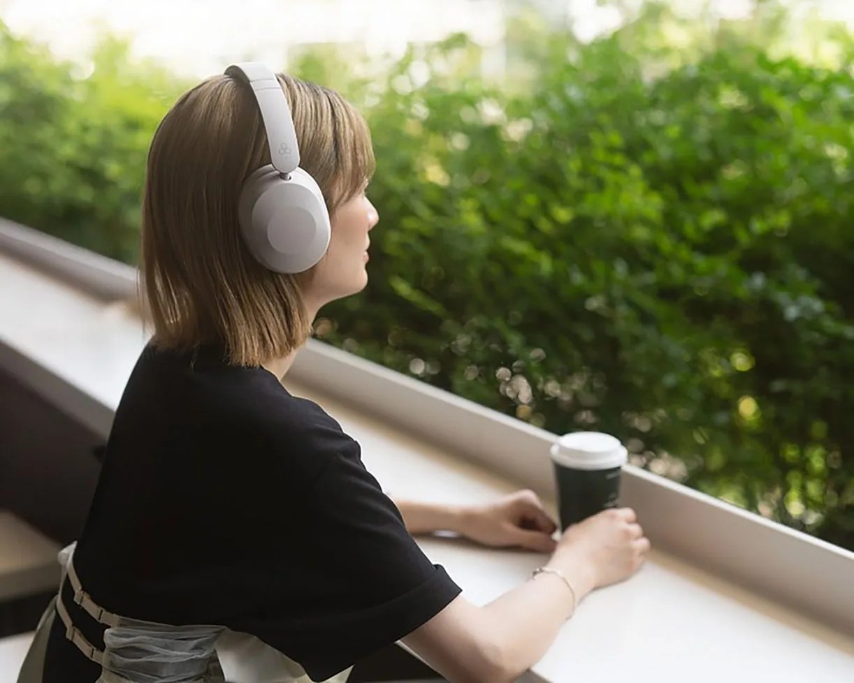 Person wearing white over-ear headphones and black shirt sitting at a white counter holding a black coffee cup with greenery outside.