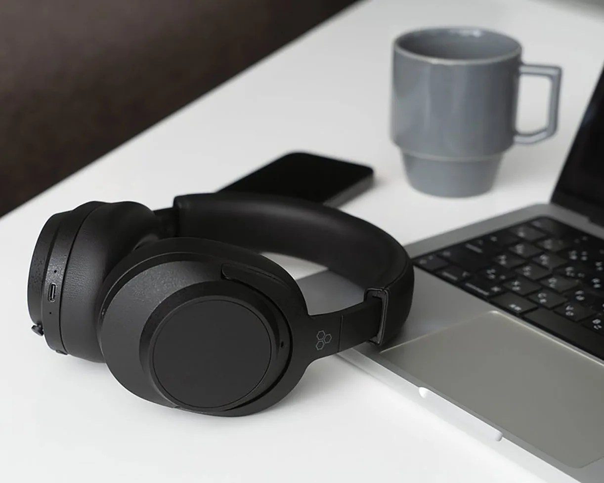 Black over-ear wireless headphones resting on a silver laptop keyboard on a white desk with a gray mug and smartphone in the background.