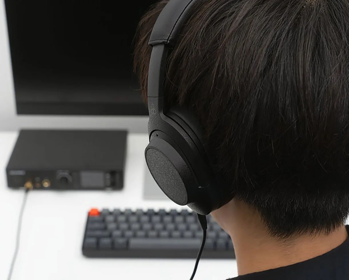 Person wearing black over-ear headphones connected to a black audio device on a white desk with a keyboard and monitor.