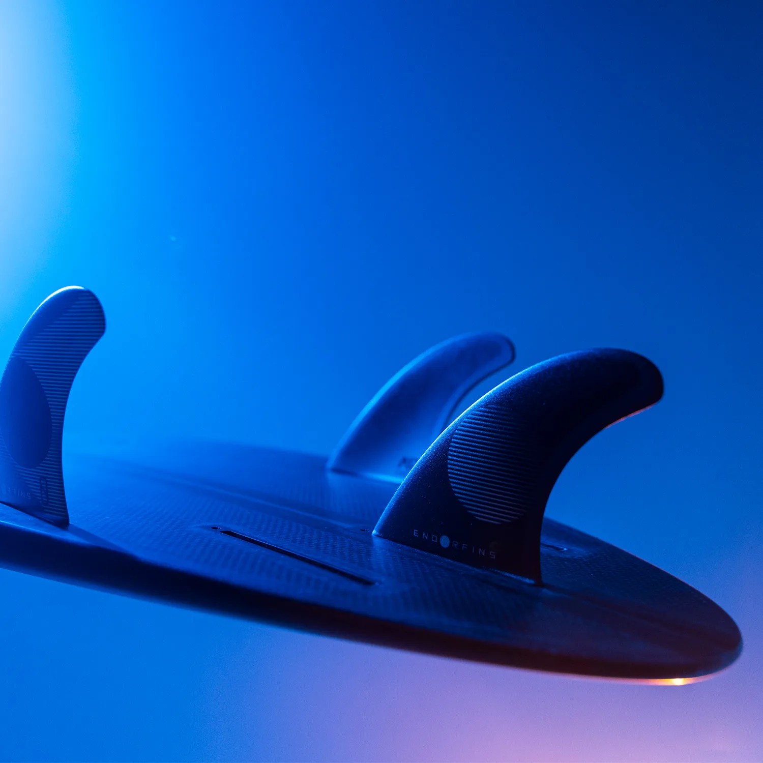 Close-up of a black surfboard with three fins under blue lighting.