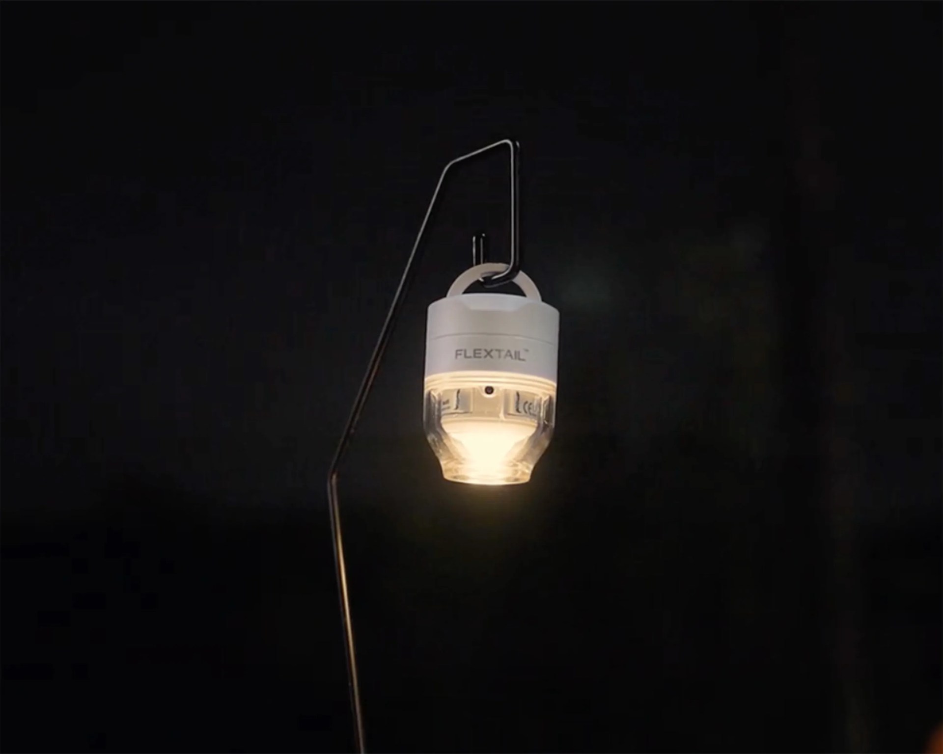 Small white and clear hanging LED lantern labeled "FLEXTAIL" glowing in the dark on a black metal stand.