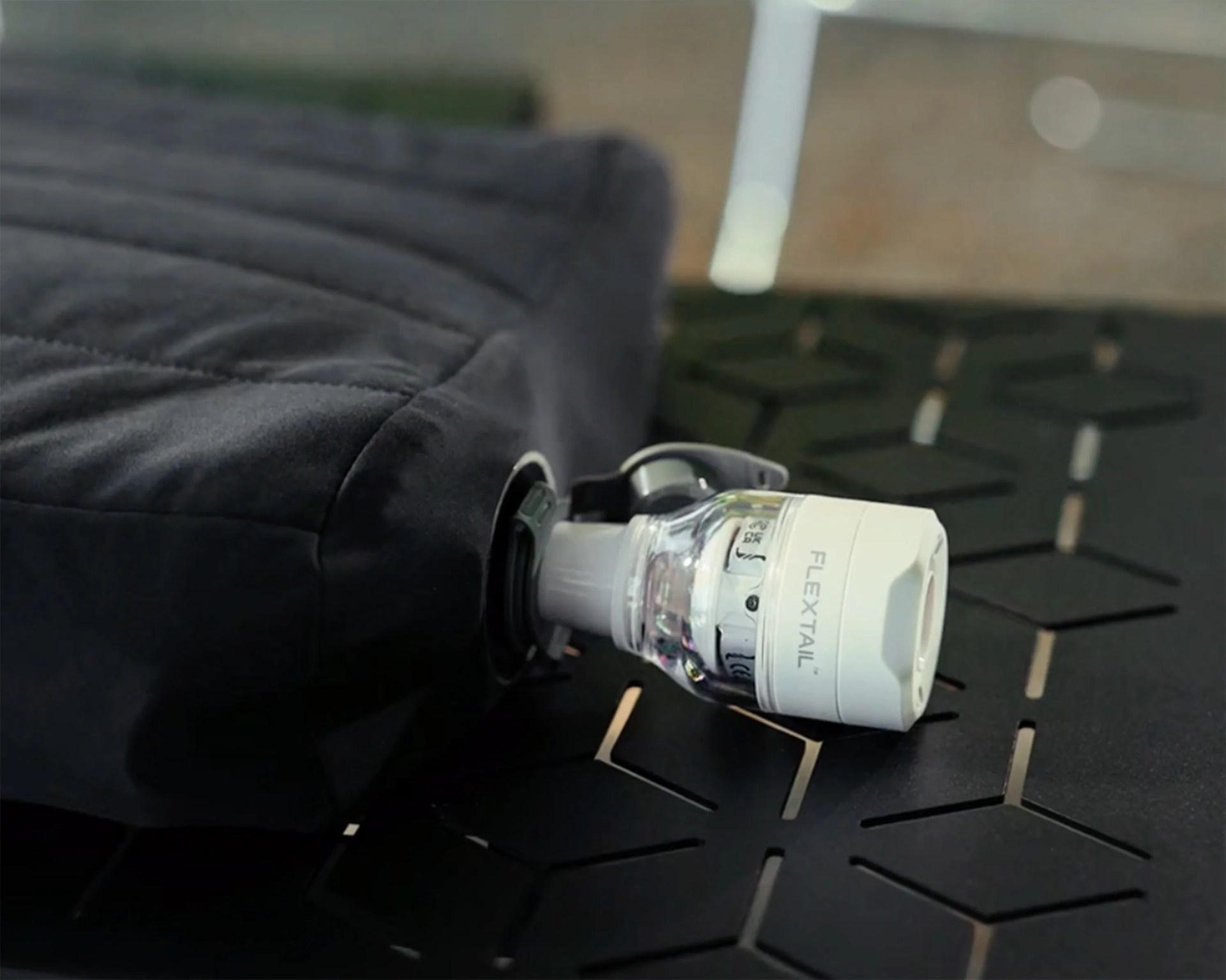 White and clear FLEXTAIL valve attached to a black fabric item on a black surface with geometric cutouts.