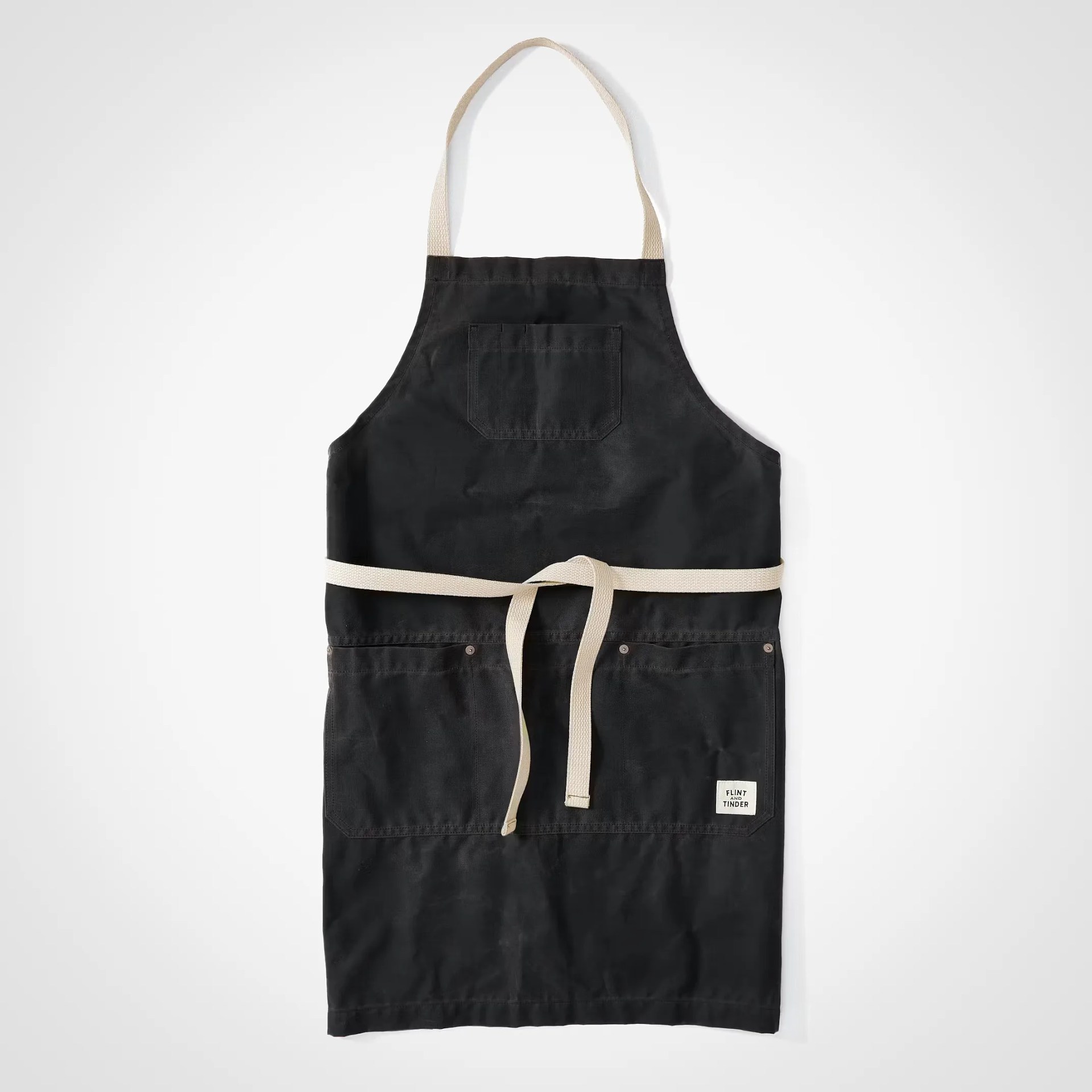 Black canvas apron with beige neck strap and waist ties, featuring front pockets and a small label.
