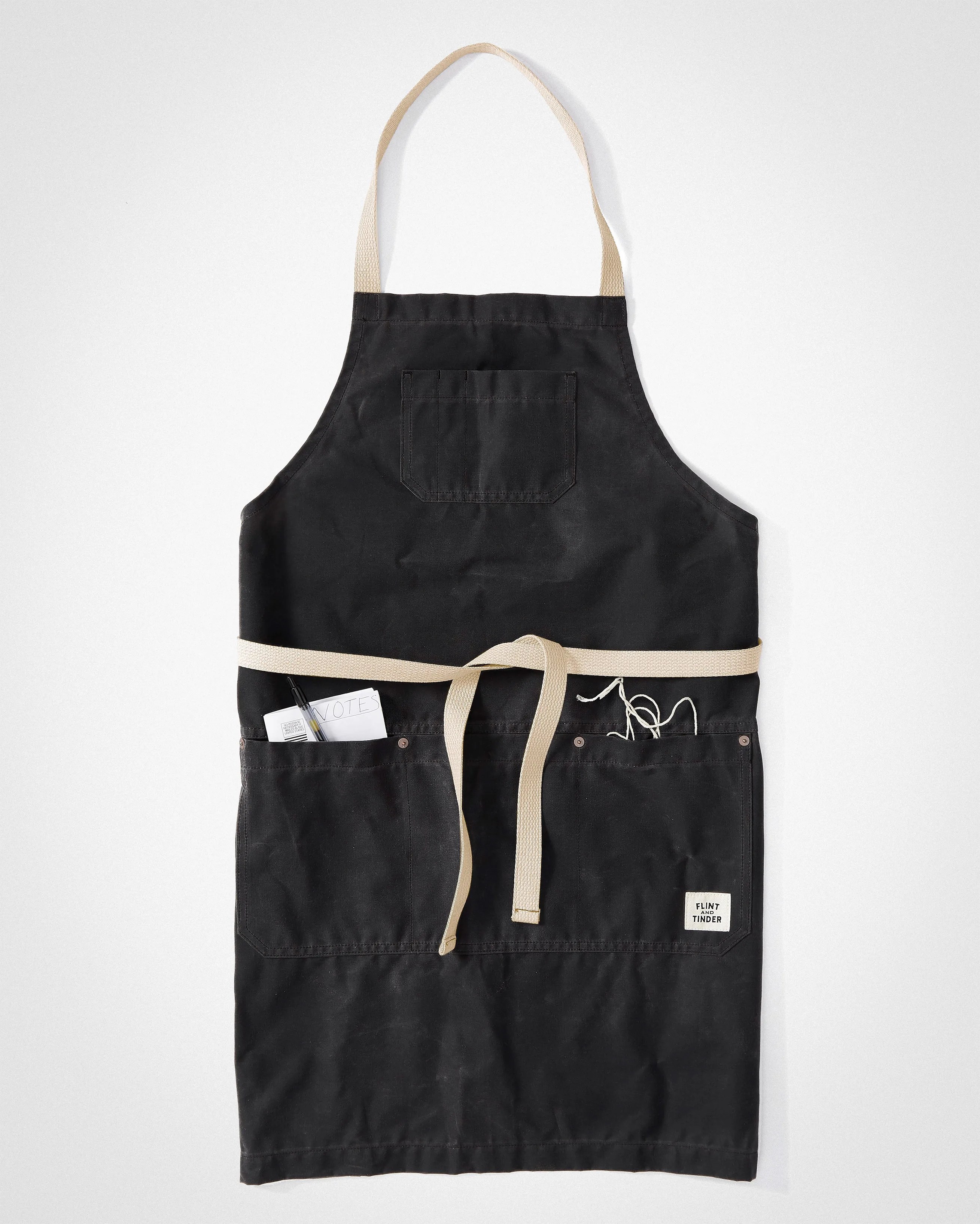 Black canvas apron with beige neck strap and waist ties, featuring front pockets and a small "Flint and Tinder" label.
