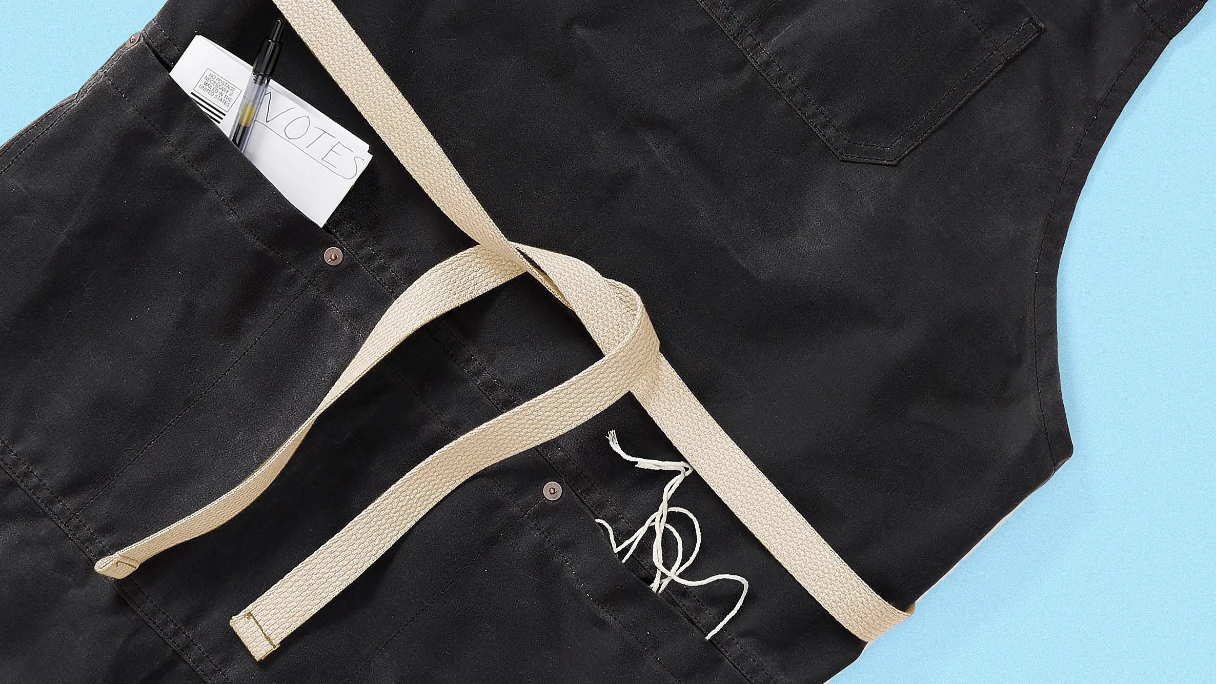 Black apron with beige straps and front pockets, one holding a pen and paper labeled "VOTES.