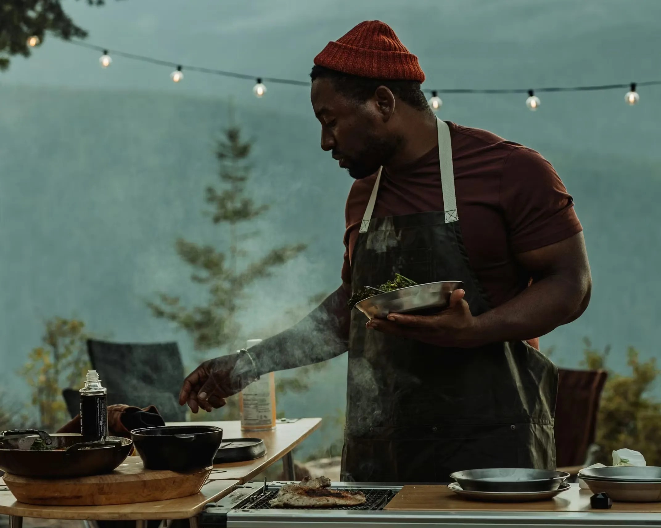 Man wearing a red beanie and apron grilling food outdoors with string lights in the background.