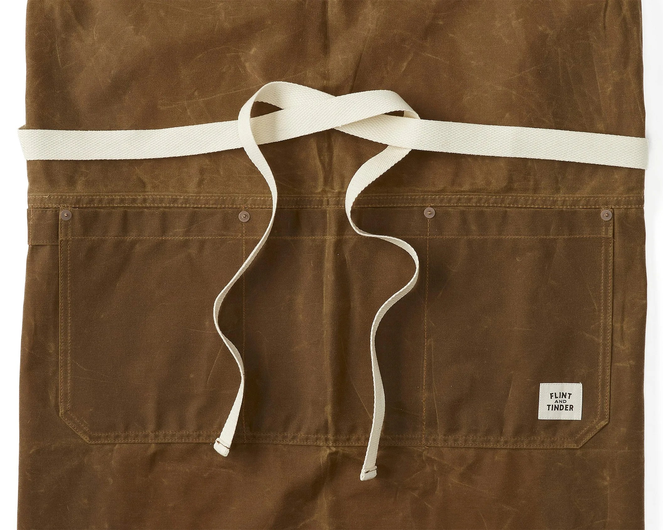 Brown waxed canvas apron with two front pockets and white waist ties, featuring a "Flint and Tinder" label.