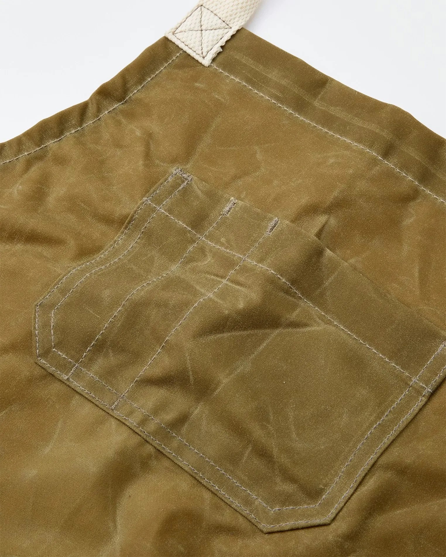 Close-up of a brown waxed canvas apron pocket with stitched compartments and a beige strap loop.