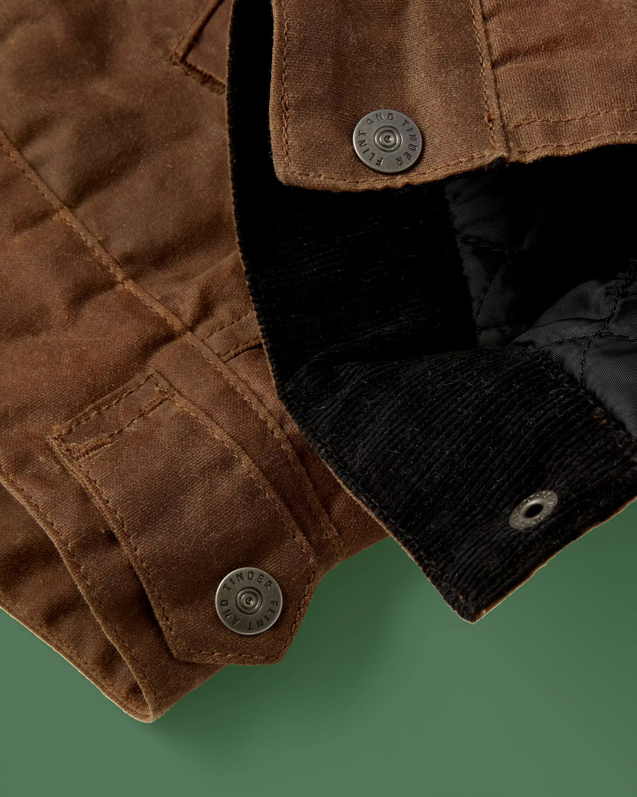 Close-up of a brown waxed cotton jacket cuff with black corduroy lining and metal snap buttons engraved with "Flint and Tinder.